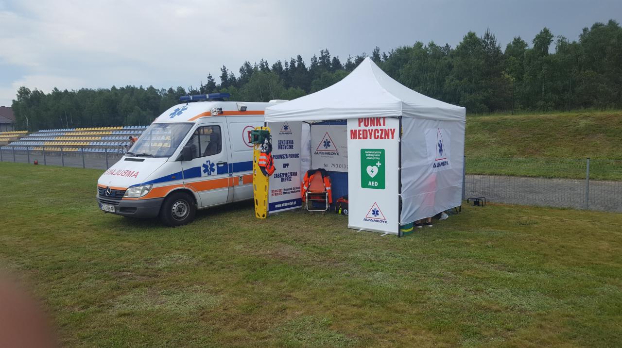 Ambulans i namiot punktu medycznego AlfaMedyk na trawiastym terenie, widoczna deska ortopedyczna i krzesełko ratownicze, w tle trybuny i las.