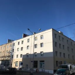 Wielopiętrowy budynek mieszkalny z nową, dwukolorową elewacją, widoczny z poziomu ulicy w słoneczny dzień, z zaparkowanymi samochodami i elementami rusztowania w tle.