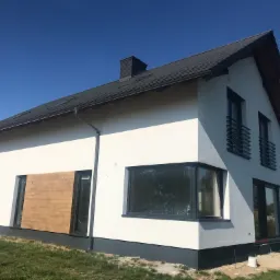 Nowoczesny dom jednorodzinny z białymi ścianami i ciemną dachówką, z częściowo zasłoniętymi oknami płytami OSB, na etapie wykończenia elewacji, widoczne materiały budowlane obok wejścia.