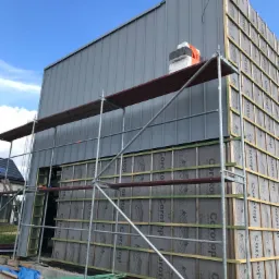 Budowa domu w trakcie ocieplania: szkielet elewacji z widoczną membraną Corotop, metalowe panele ścienne, rusztowanie i niebieskie niebo w tle.