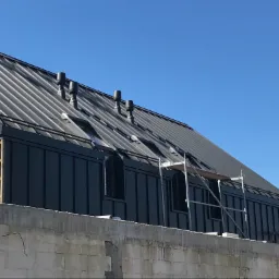 Szara blacha na dachu z trzema kominami wentylacyjnymi, widok z dołu na budynek w stanie surowym w Łodzi, z widocznym rusztowaniem.