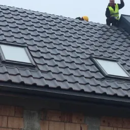 Ekipa dekarzy na dachu z dachówką ceramiczną, widoczne dwa okna dachowe, jeden pracownik trzyma kask w górze, budynek w stanie surowym otwartym.