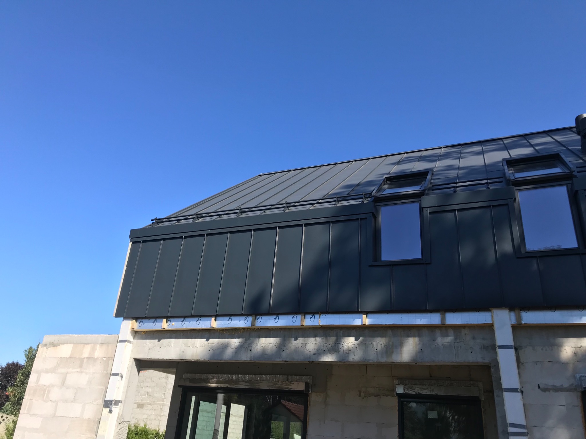 Nowoczesny dom w trakcie budowy z ciemnoszarą blachą na dachu i oknami dachowymi, widoczne materiały izolacyjne na ścianach, błękitne niebo w tle.