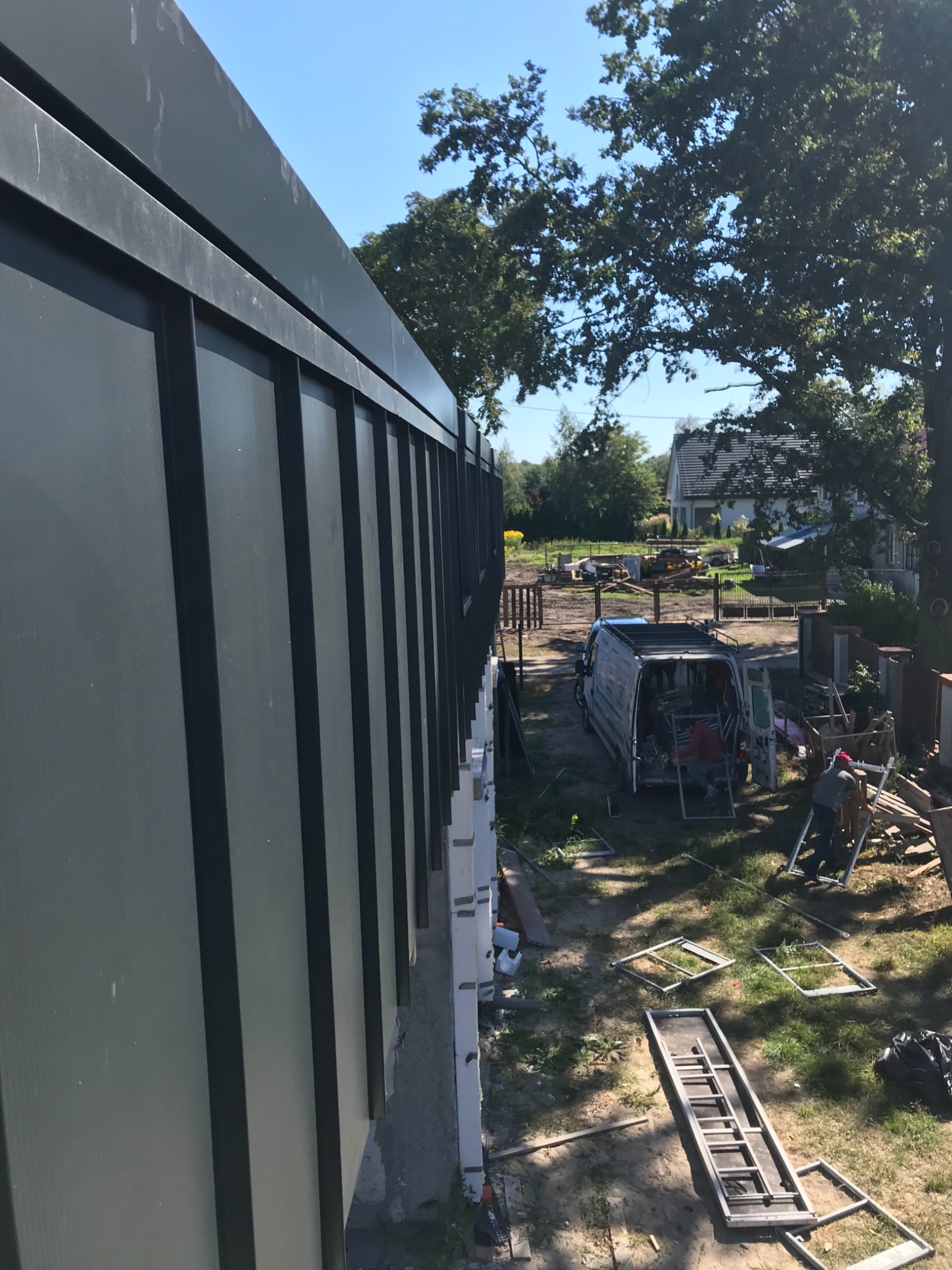 Montaż elewacji z paneli pionowych w kolorze grafitowym, widoczny samochód dostawczy z narzędziami i pracownicy w tle.