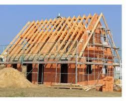 Budowa domu jednorodzinnego w trakcie wznoszenia: widoczna konstrukcja ścian z czerwonej cegły, drewniana konstrukcja dachu w trakcie montażu, rusztowania, hałda piasku i paleta cegieł na pierwszym...