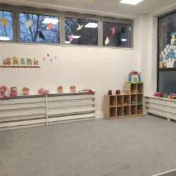 Jasne, przestronne wnętrze żłobka z szarym dywanem, białymi ścianami ozdobionymi naklejkami z motywem pociągu i balonów, oraz kolorowymi ptakami w oknach. Na grzejniku ustawione lalki, a obok...