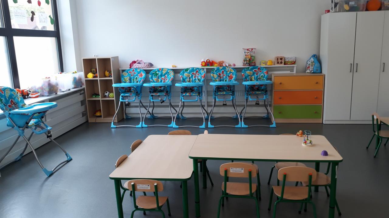 Jasne wnętrze sali żłobkowej z rzędem niebieskich krzesełek do karmienia, stolikami i krzesłami dla dzieci oraz regałami z zabawkami.