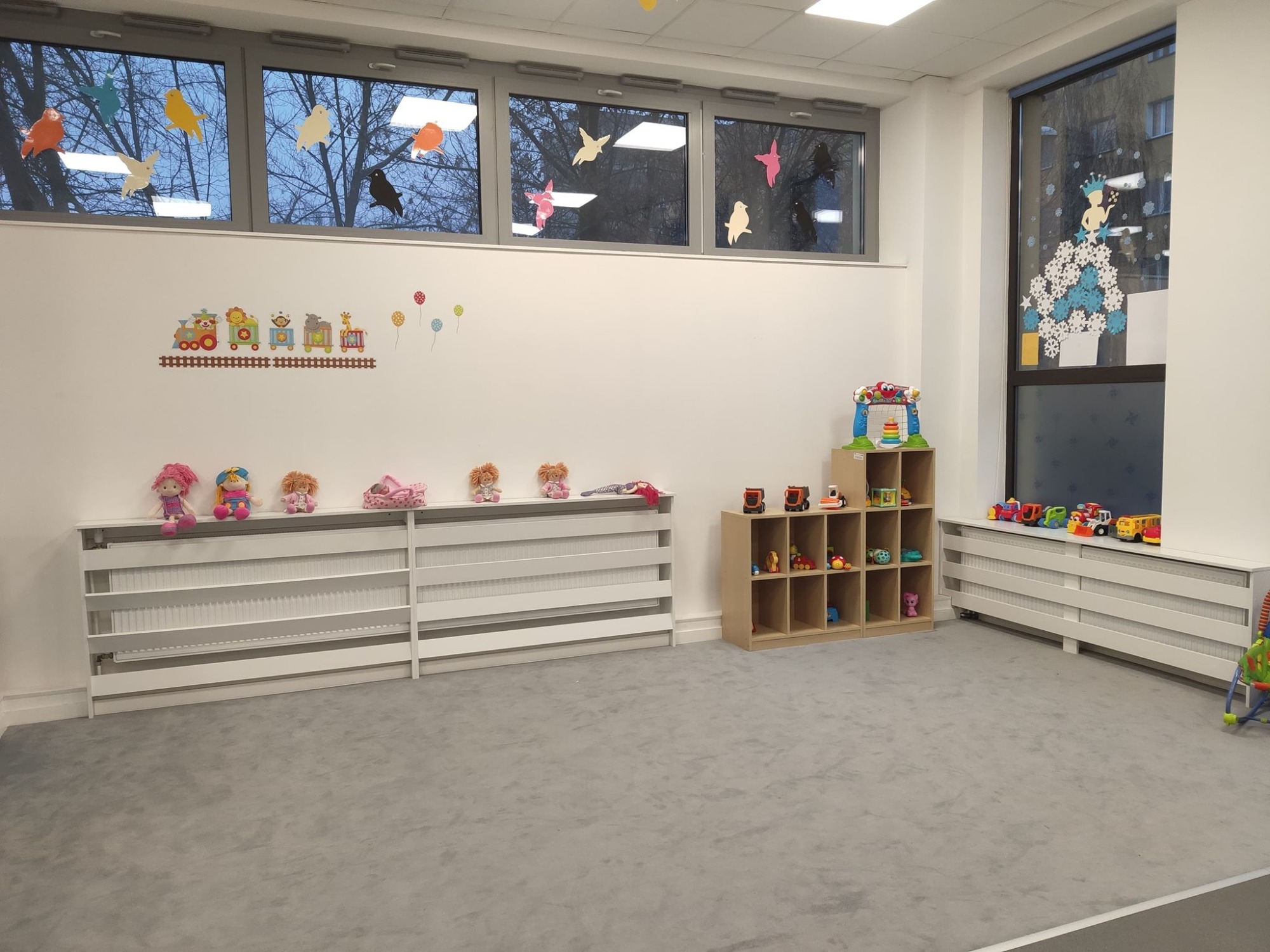 Jasne, przestronne wnętrze żłobka z szarym dywanem, białymi ścianami ozdobionymi naklejkami z motywem pociągu i balonów, oraz kolorowymi ptakami w oknach. Na grzejniku ustawione lalki, a obok...