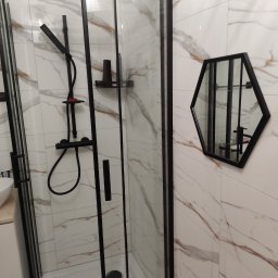 Sław-Bud Firma remontowo-budowlana - Kabina prysznicowa z czarnymi ramami i armaturą, heksagonalne lustro na ścianie z marmurowym wzorem.