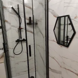 Kabina prysznicowa z czarnymi ramami i armaturą, heksagonalne lustro na ścianie z marmurowym wzorem.