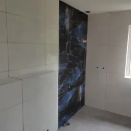 Wnętrze łazienki w trakcie prac wykończeniowych: białe płytki ścienne, fragment ściany z ciemnymi płytkami imitującymi marmur, okno z widokiem na zewnątrz.