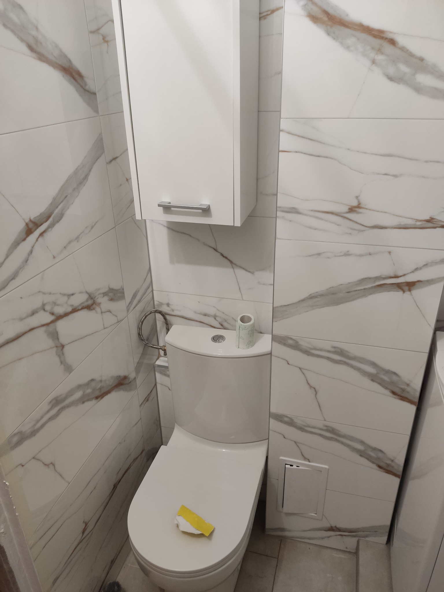 Wykończona toaleta z białymi marmurkowymi płytkami, wiszącą szafką i rolką papieru toaletowego na spłuczce; widoczny fragment żółtej taśmy malarskiej na desce sedesowej.