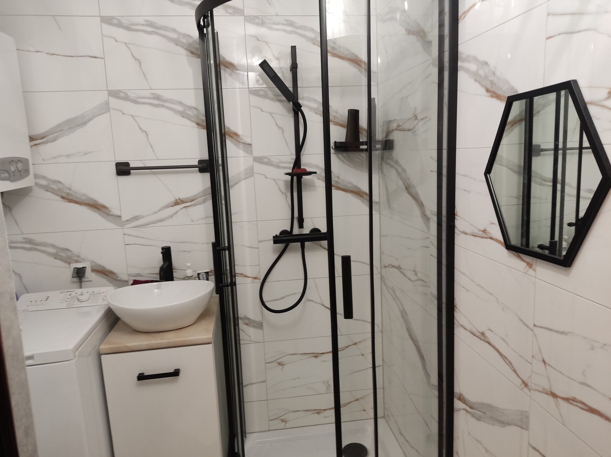 Nowoczesna łazienka z kabiną prysznicową z czarnymi ramami, umywalką nablatową i heksagonalnym lustrem na ścianie wyłożonej białymi płytkami imitującymi marmur.