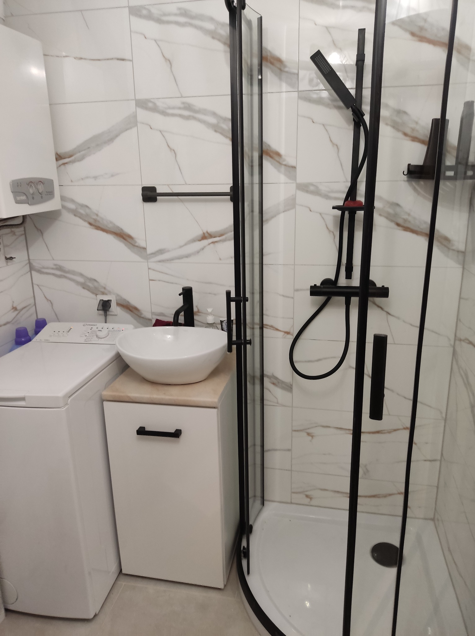Nowoczesna łazienka z kabiną prysznicową z czarnymi profilami, umywalką nablatową na szafce i pralką obok, wykończona płytkami imitującymi marmur.