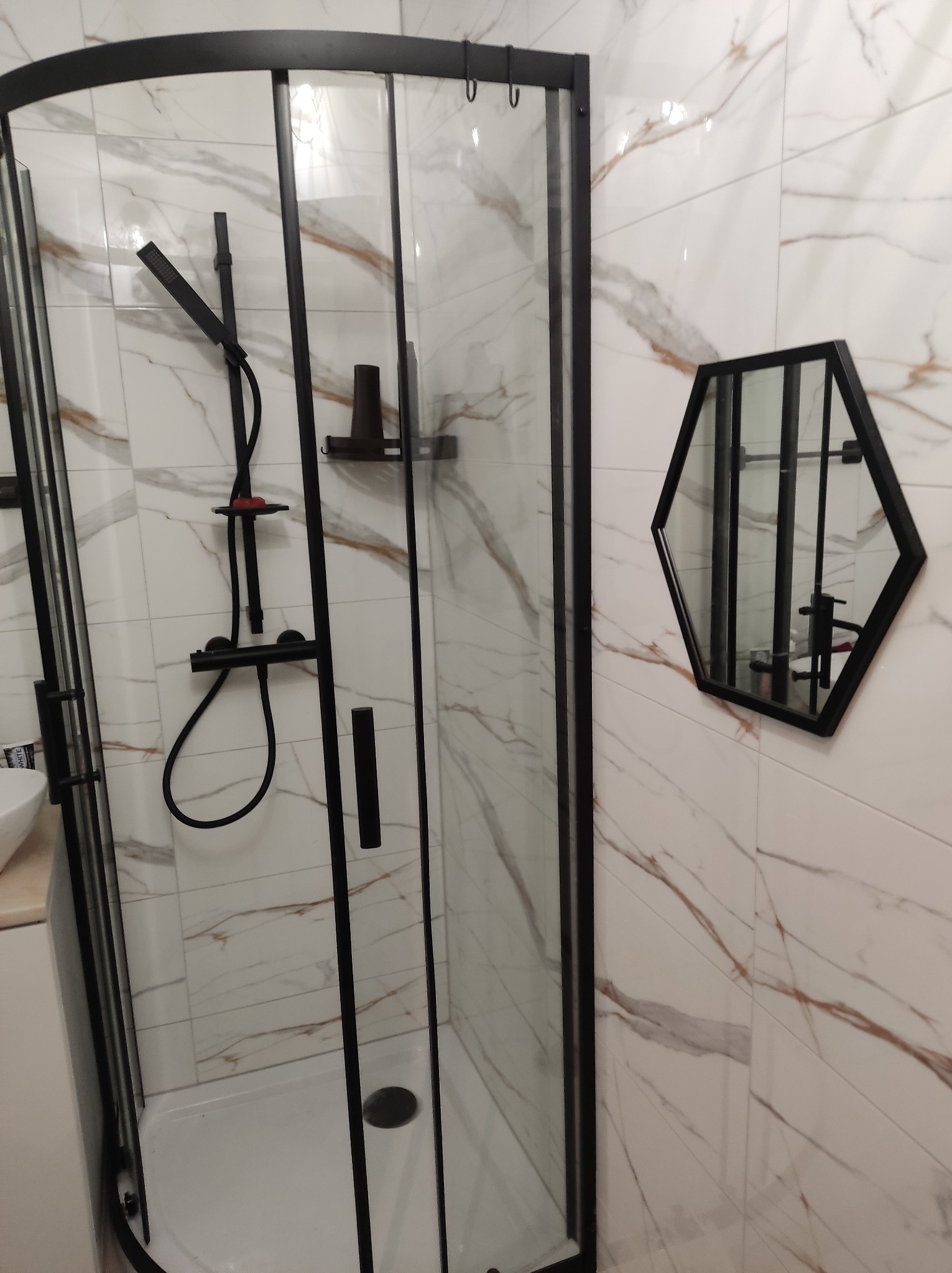 Kabina prysznicowa z czarnymi ramami i armaturą, heksagonalne lustro na ścianie z marmurowym wzorem.