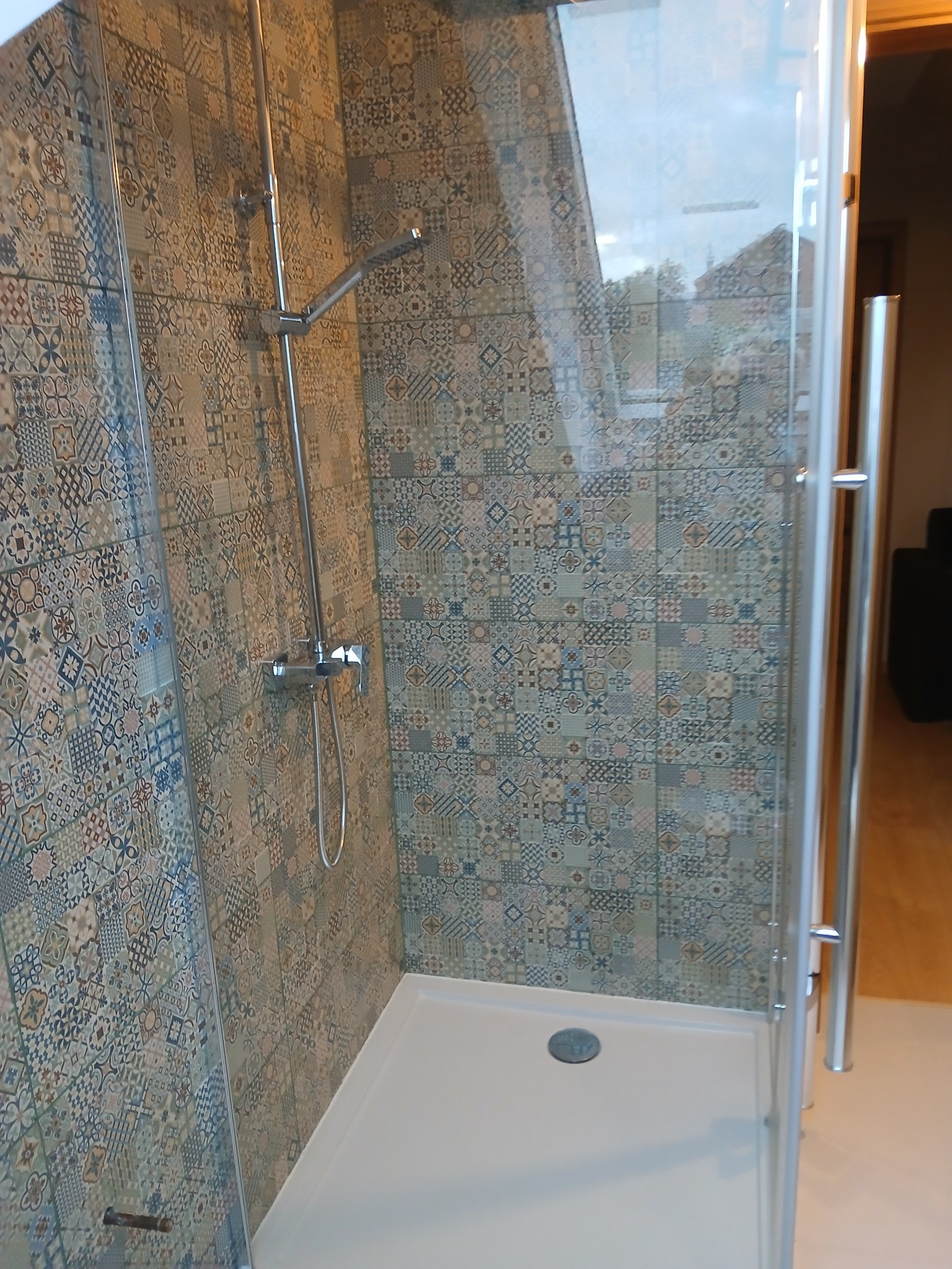 Kabina prysznicowa z mozaiką w łazience. Biały brodzik, chromowana armatura. Widok na wzorzyste płytki i szklane drzwi kabiny.