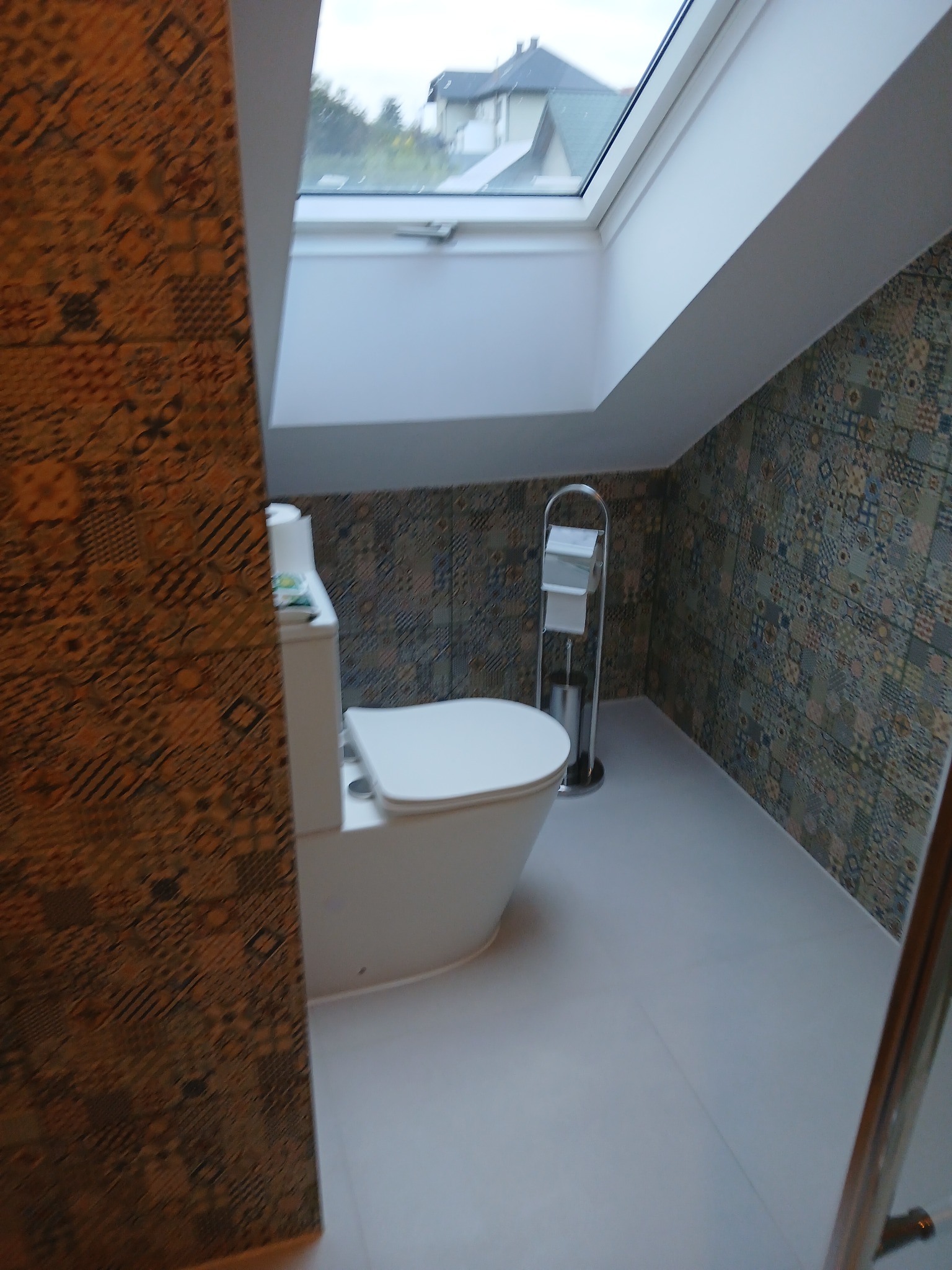 Wykończona łazienka na poddaszu z oknem dachowym. Uwagę przyciągają wzorzyste płytki na ścianach i zabudowa WC. Widoczny również stojak na papier toaletowy i szczotkę.