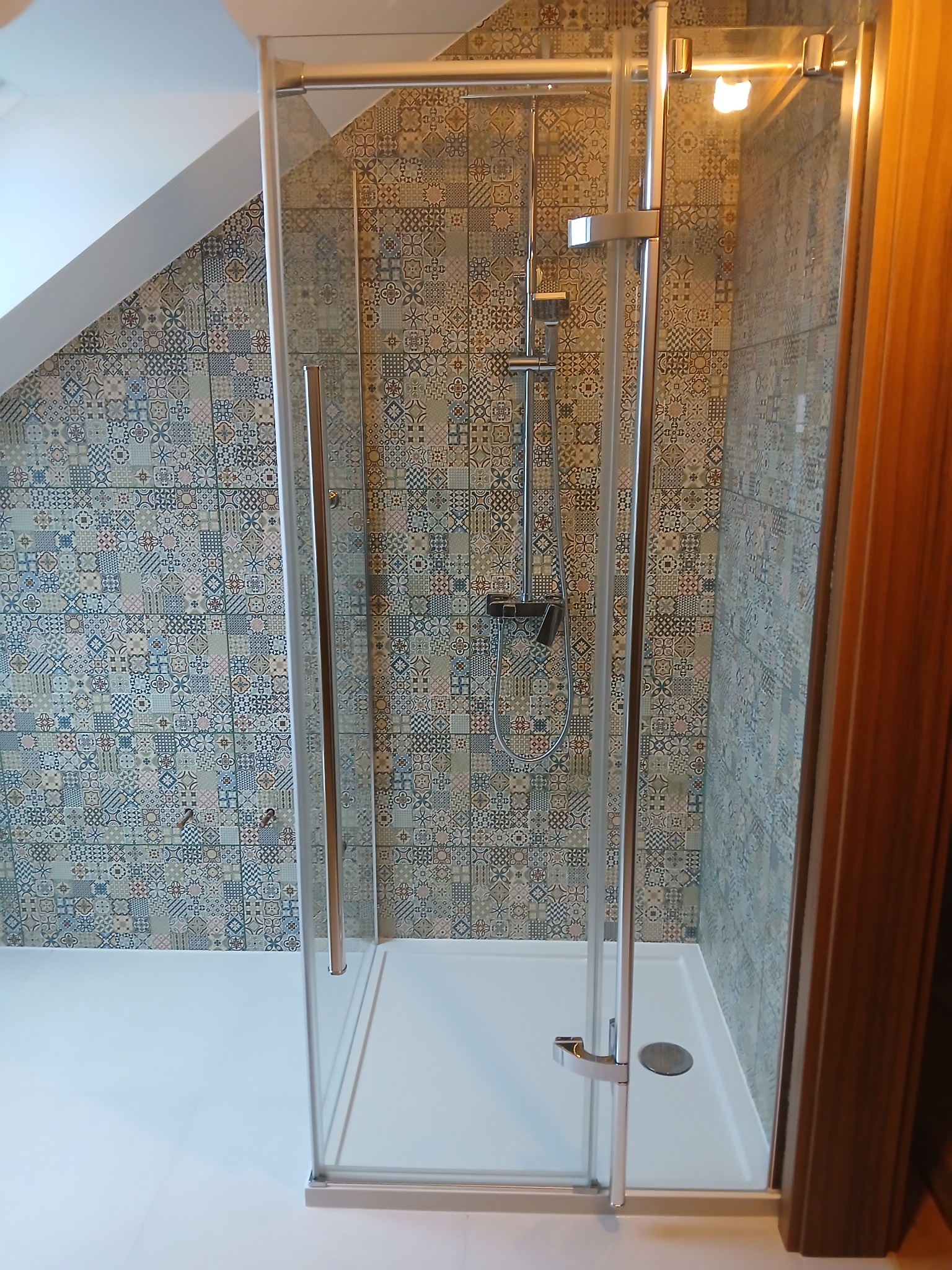 Kabina prysznicowa z mozaiką na ścianie w łazience na poddaszu. Szklane drzwi z srebrnym uchwytem. Biały brodzik i podłoga. Nowoczesny design.