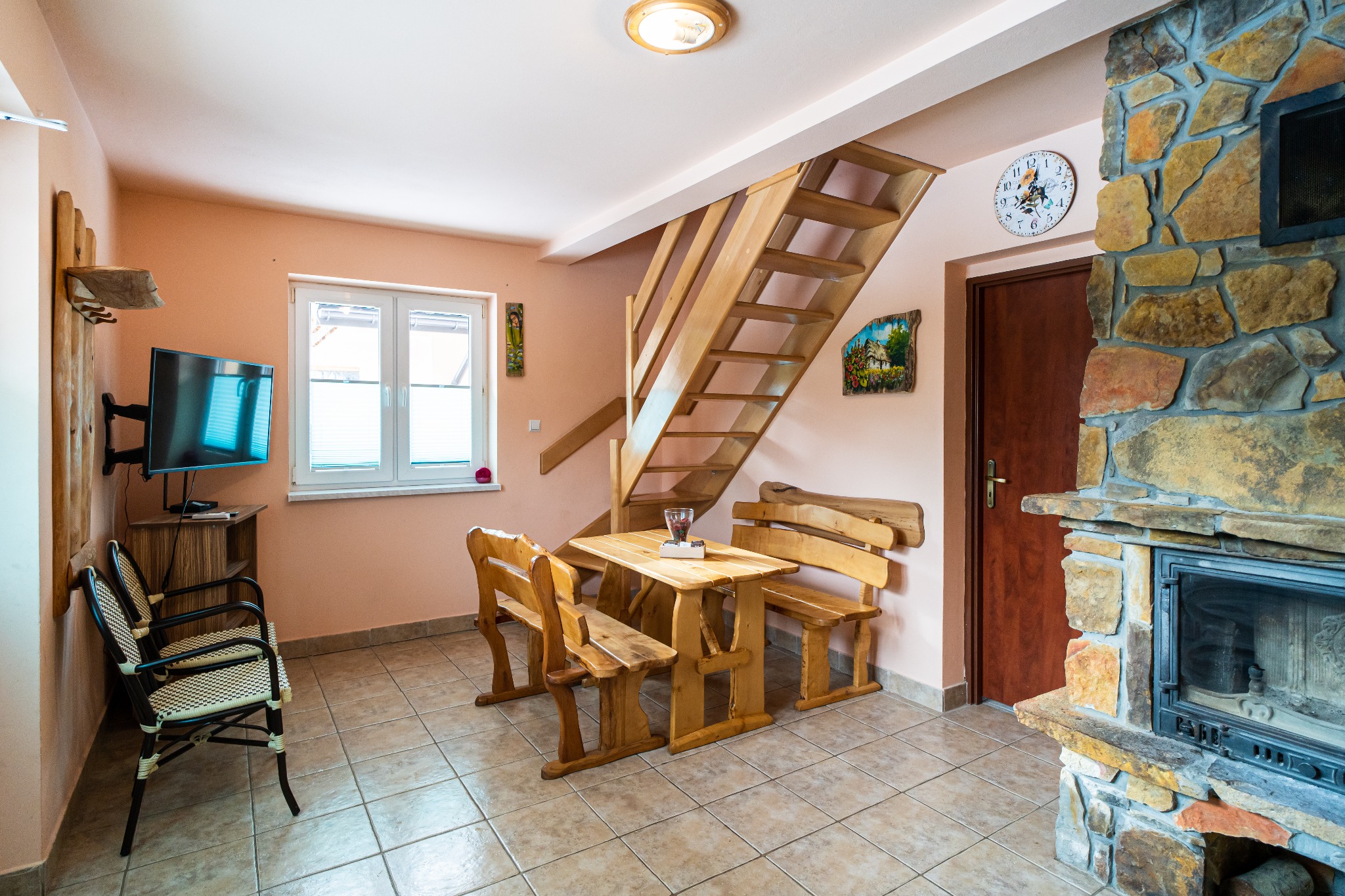 Rustykalne wnętrze pokoju z drewnianym stołem, ławami, schodami i kamiennym kominkiem, oświetlone ciepłym światłem.