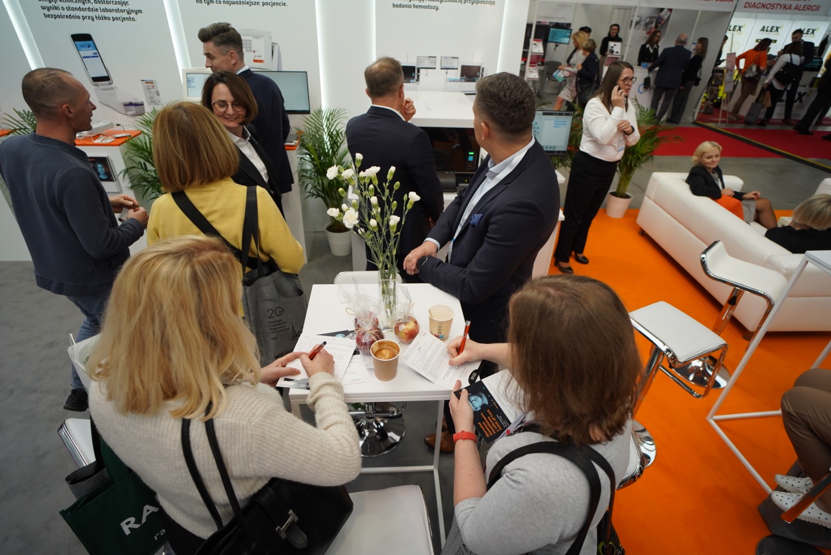 Osoby wypełniają formularze przy stoliku na targach, w tle stanowisko z wyposażeniem medycznym i roślinami, widoczna kawa i jabłka.