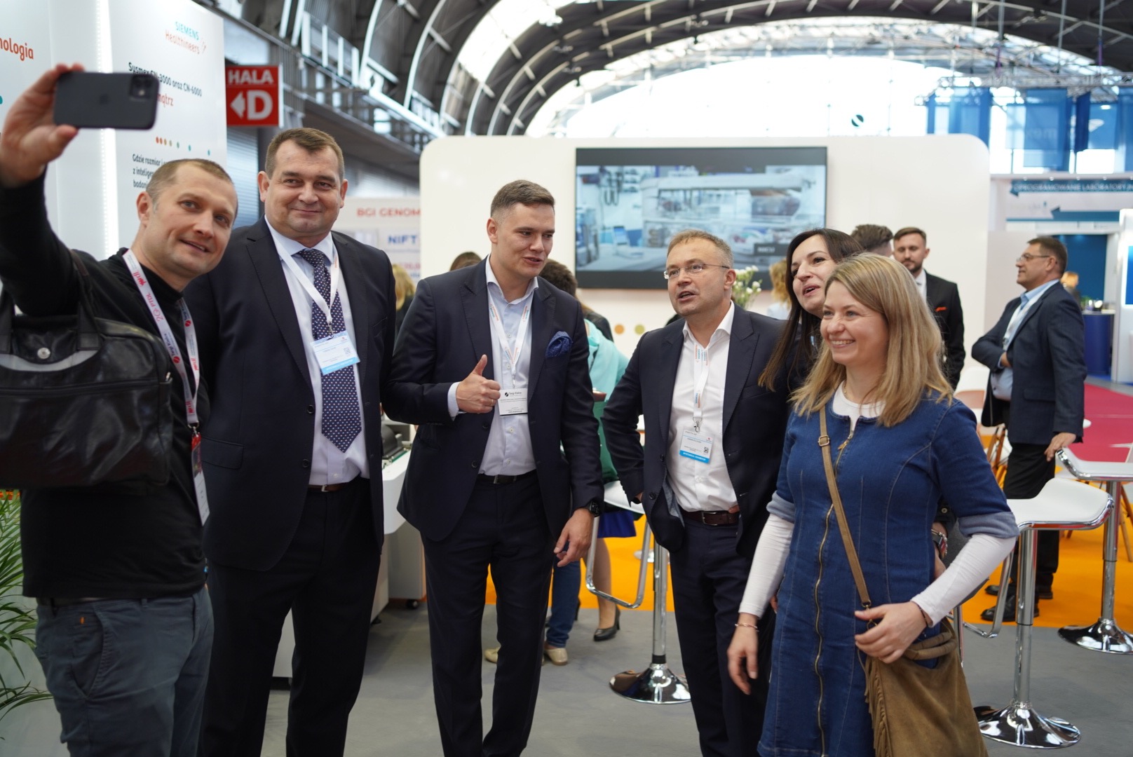 Grupa osób, w tym mężczyźni w garniturach i kobieta w dżinsowej sukience, pozuje do zdjęcia na tle stoiska targowego z logo firm i ekranem prezentującym sprzęt laboratoryjny.