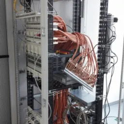 Otwarta szafa serwerowa z licznymi kablami pomarańczowymi, patch panele i urządzenia sieciowe, widoczne narzędzia (śrubokręt) na półce, nieuporządkowane okablowanie na dole szafy.