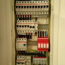 Otwarta skrzynka elektryczna z bezpiecznikami, listwami zaciskowymi, gniazdem i przewodami w kolorach: zielono-żółtym, niebieskim i czarnym. Widoczne zabezpieczenia przeciwprzepięciowe w kolorze...