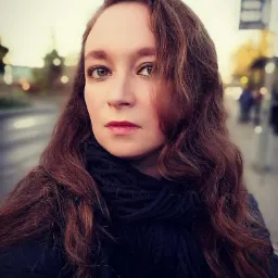 Portret kobiety z długimi, brązowymi włosami i ciemnym szalem na tle miejskiego krajobrazu w rozmyciu, z widocznymi elementami infrastruktury miejskiej.