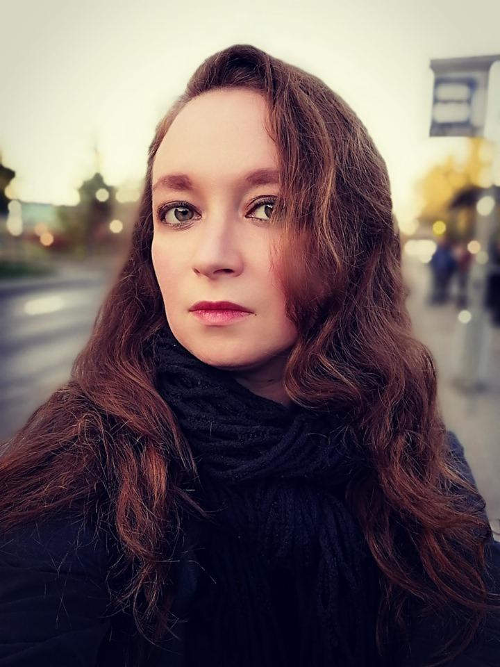 Portret kobiety z długimi, brązowymi włosami i ciemnym szalem na tle miejskiego krajobrazu w rozmyciu, z widocznymi elementami infrastruktury miejskiej.