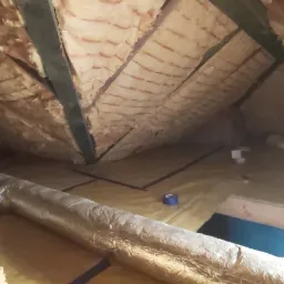 Izolacja poddasza wełną mineralną między krokwiami, folia paroizolacyjna na podłodze, widoczny przewód wentylacyjny w folii aluminiowej i niebieska taśma malarska.