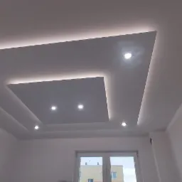 Sufit podwieszany z oświetleniem LED w kształcie kwadratów, widoczne profile metalowe na krawędziach sufitu, okno w tle.