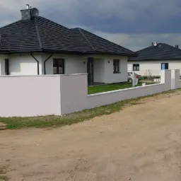 Nowoczesne domy jednorodzinne z ciemnymi dachami i jasnymi elewacjami, ogrodzone niskim, pomalowanym murem, widoczne ślady prac malarskich na murze i chodniku, osoba pracująca przy murze, samochód...