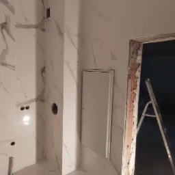 Wnętrze łazienki w Chojne w trakcie remontu: białe płytki imitujące marmur na ścianach i podłodze, widoczne otwory na instalacje, drabina oparta o futrynę drzwi.