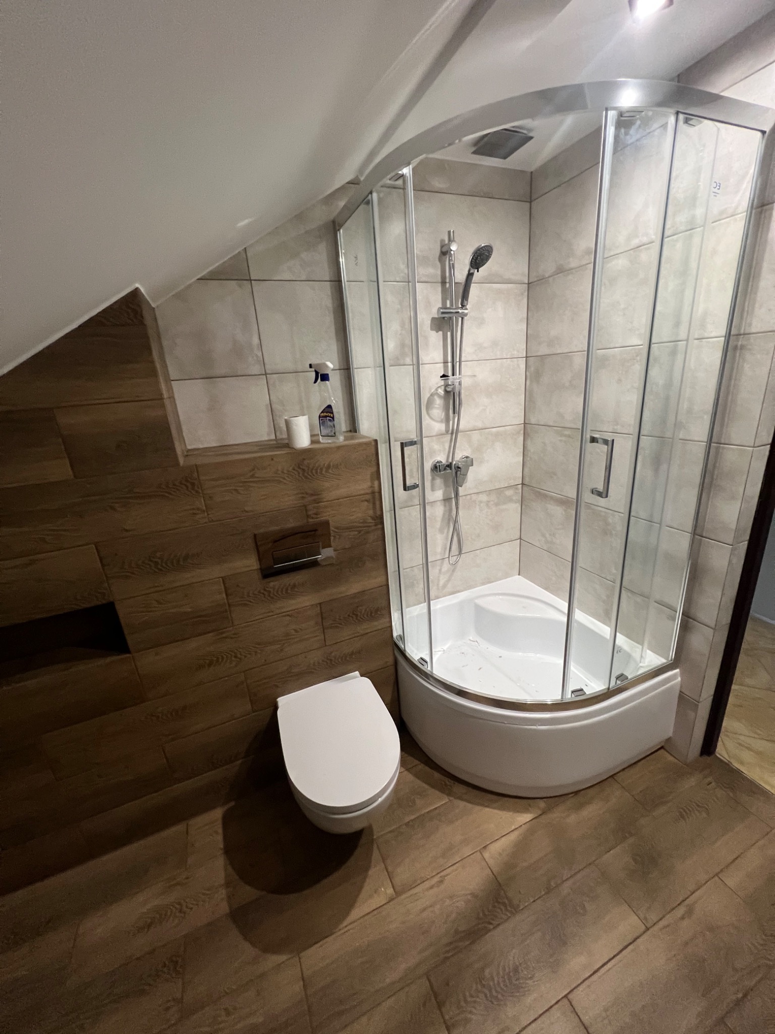 Ukończona łazienka na poddaszu ze skośnym sufitem: kabina prysznicowa narożna z brodzikiem, podwieszana toaleta i drewnopodobne płytki na ścianach i podłodze.