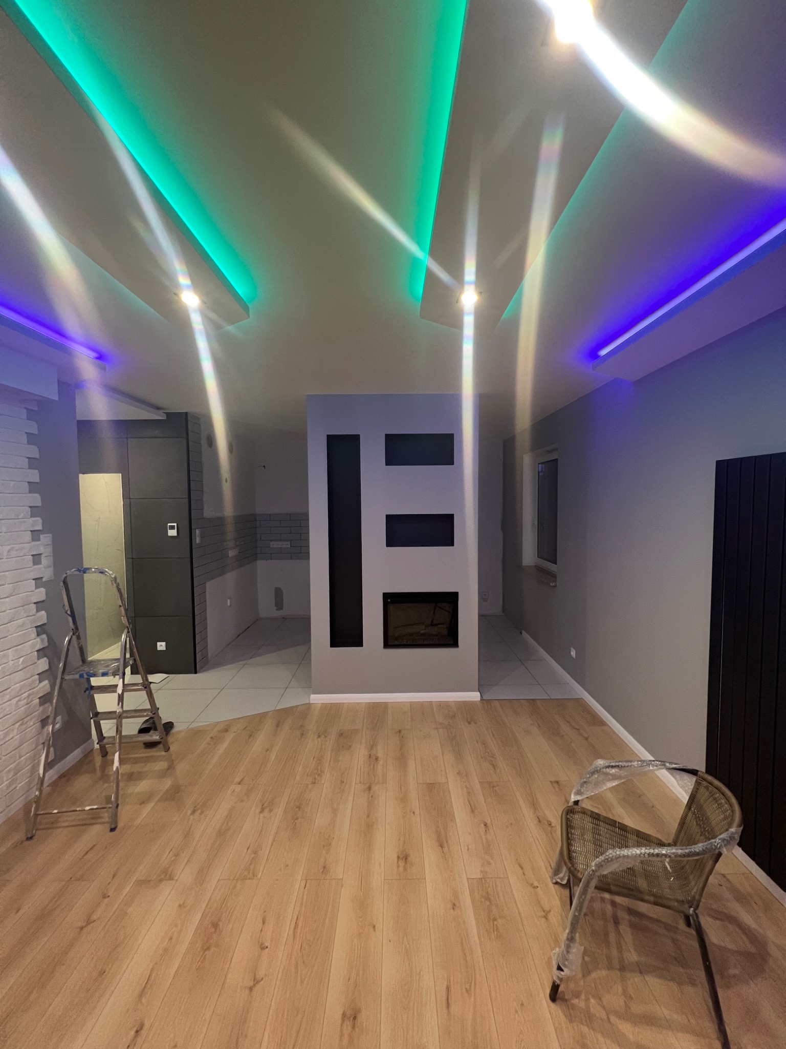 Wnętrze w trakcie prac wykończeniowych: drewnopodobna podłoga, ściany pomalowane na szaro, dekoracyjny kominek z czarnymi wstawkami, schody malarskie i krzesło zabezpieczone folią, sufit...