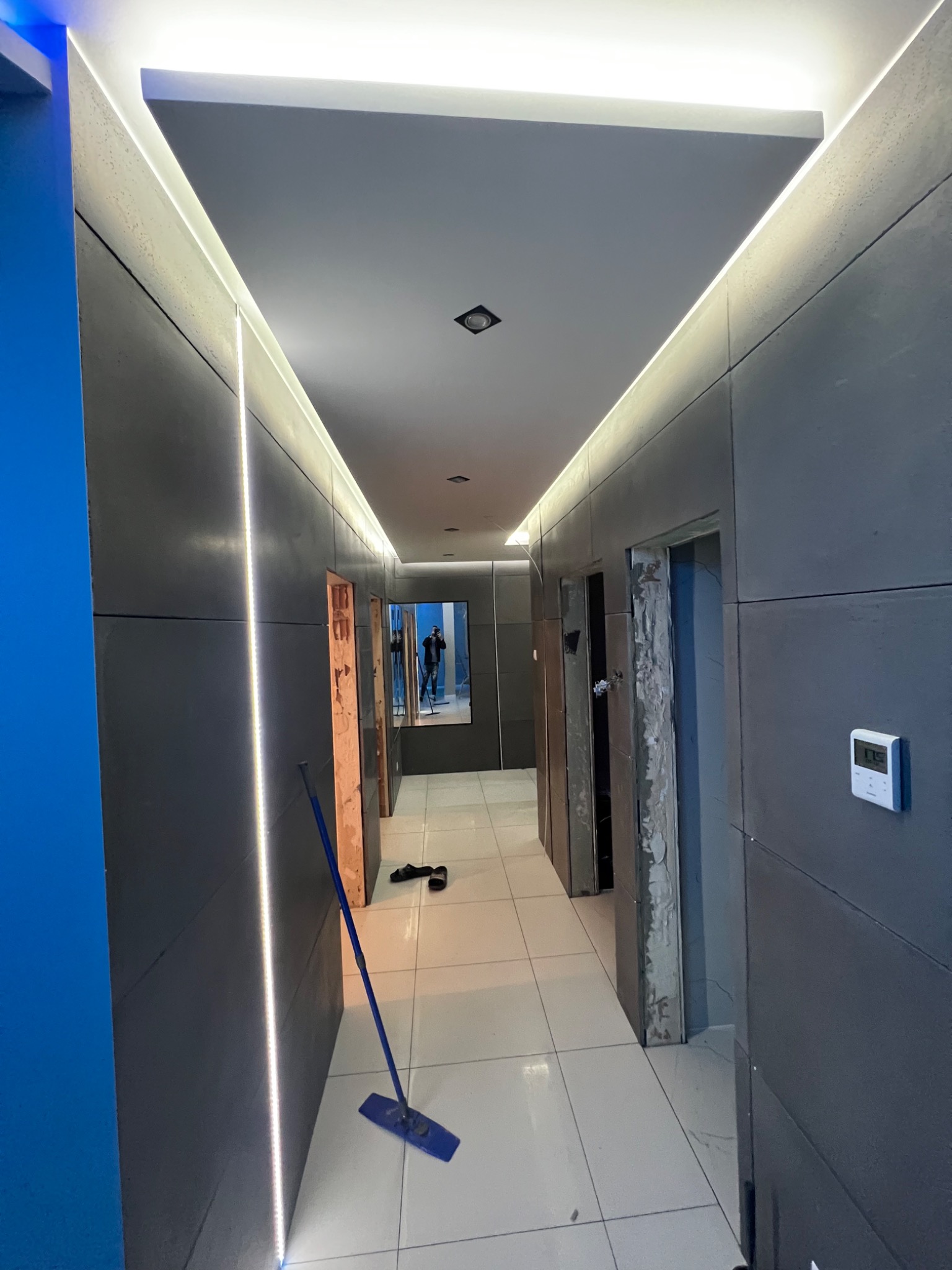 Korytarz w trakcie wykańczania z szarymi ścianami, podświetlanym sufitem i podłogą z płytek, widoczne otwarte ościeżnice drzwiowe bez drzwi, mop stojący na podłodze.