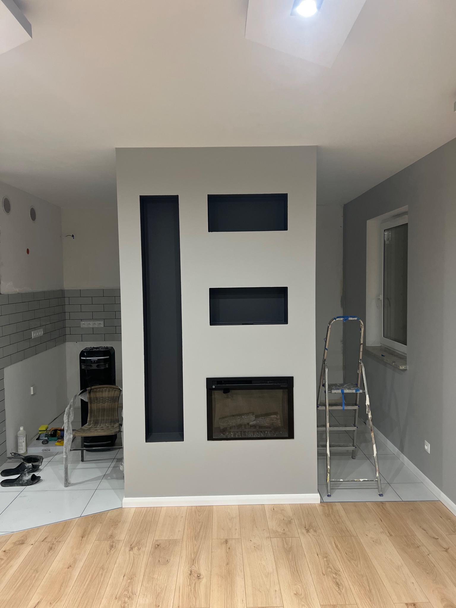 Wnętrze w Chojnem w trakcie prac wykończeniowych: ściana z zabudową kominkową z wnękami pomalowana na szaro, widoczna drabina malarska, fragment podłogi wyłożonej panelami oraz fragment ściany...