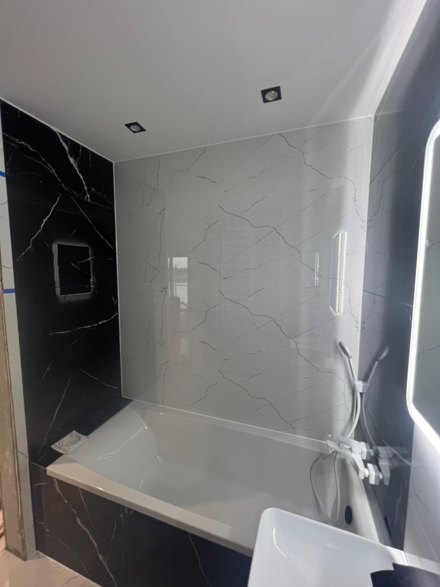 Wykończona łazienka z wanną obłożoną czarno-białymi płytkami imitującymi marmur, widoczna armatura i oświetlenie punktowe.