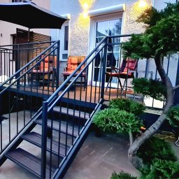 Artifex Radosław Stąsiek - Metalowe schody z balustradą prowadzące na taras z meblami ogrodowymi i oświetleniem, otoczone zielenią. Nowoczesny design z elementami dekoracyjnymi.