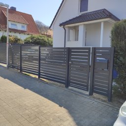 Artifex Radosław Stąsiek - Nowoczesne, poziome ogrodzenie panelowe w kolorze antracytowym, z furtką i skrzynką na listy, na tle domu z dwuspadowym dachem i kostki brukowej.