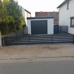 Nowoczesne ogrodzenie panelowe poziome w kolorze antracytowym, z bramą wjazdową i furtką, otaczające posesję z garażem i domem w tle.