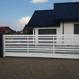 Nowoczesna, przesuwna brama wjazdowa z białymi, poziomymi profilami na tle domu z ciemną dachówką i szarą nawierzchnią z kamyków.