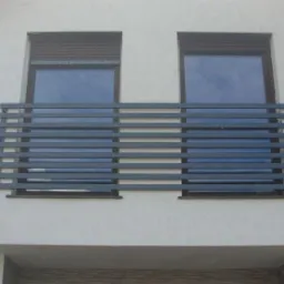 Szara, pozioma balustrada okienna z profili zamkniętych na elewacji domu, z roletami zewnętrznymi częściowo zasłoniętymi.