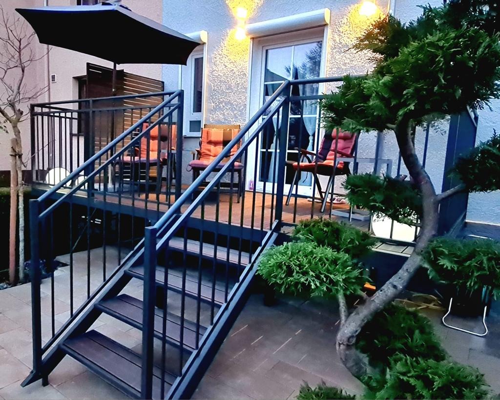 Metalowe schody z balustradą prowadzące na taras z meblami ogrodowymi i oświetleniem, otoczone zielenią. Nowoczesny design z elementami dekoracyjnymi.