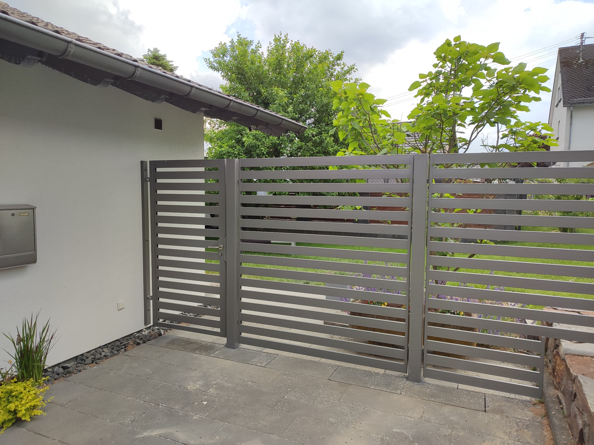 Nowoczesna, pozioma brama wjazdowa z szarych, metalowych profili, z widoczną skrzynką na listy i roślinnością u podstawy, na tle zieleni drzew i fragmentu domu.