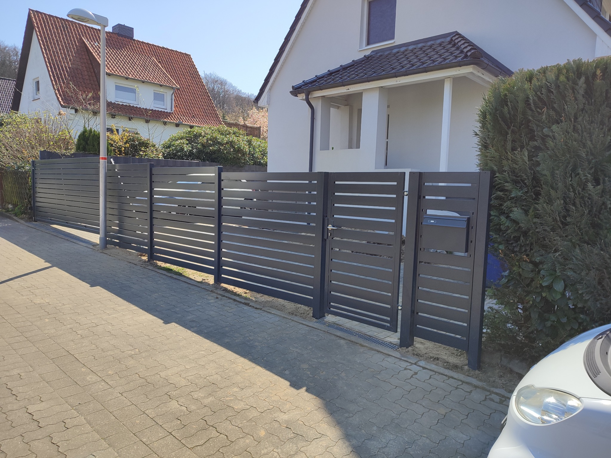 Nowoczesne, poziome ogrodzenie panelowe w kolorze antracytowym, z furtką i skrzynką na listy, na tle domu z dwuspadowym dachem i kostki brukowej.