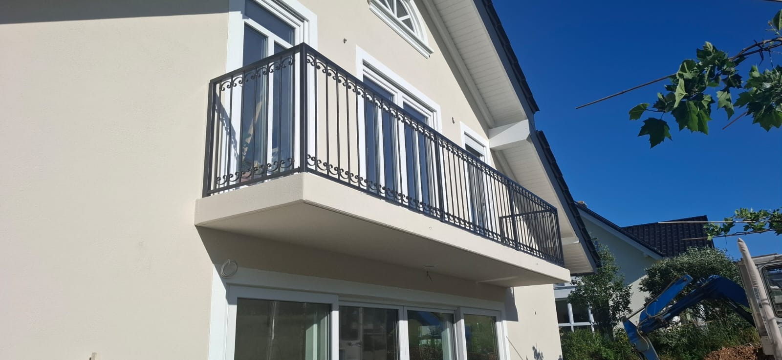 Elegancka, czarna balustrada balkonowa z dekoracyjnymi elementami, zamontowana na jasnym balkonie budynku z białymi oknami i błękitnym niebem w tle.