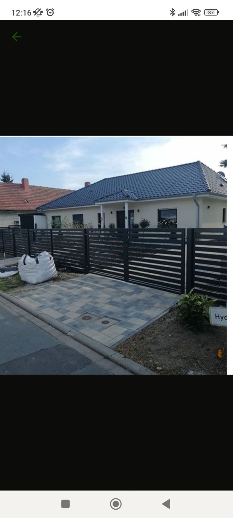 Nowoczesne ogrodzenie metalowe z poziomymi panelami w kolorze antracytowym, posesja z kostki brukowej, dom jednorodzinny w tle.