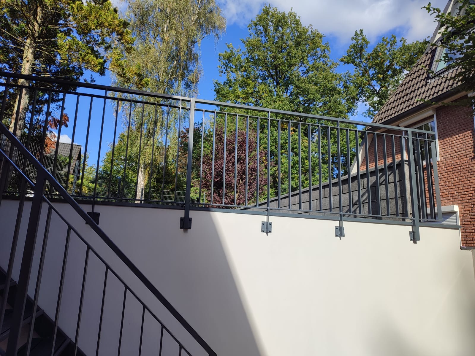 Nowoczesna, metalowa balustrada schodowa w kolorze grafitowym, z pionowymi szczeblami, zamontowana na jasnej ścianie. Widok z dołu, z fragmentem schodów i zielenią w tle.