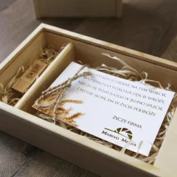 Drewniane pudełko z pendrivem 'Film Ślubny', kartką z życzeniami i logo Marivo Media, wypełnione dekoracyjną wełną drzewną, prezent na drewnianej podłodze.